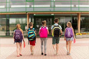 Five children wearing backpacks walking toward their elementary school.